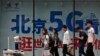FILE - People walk toward a police robot mounted with surveillance cameras patrol past a 5G network advertisement at a shopping district in Beijing, May 15, 2019. 