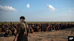 FILE - U.S.-backed Syrian Democratic Forces (SDF) fighters stand guard next to men waiting to be screened after being evacuated out of the last territory held by Islamic State group militants, near Baghouz, eastern Syria. 