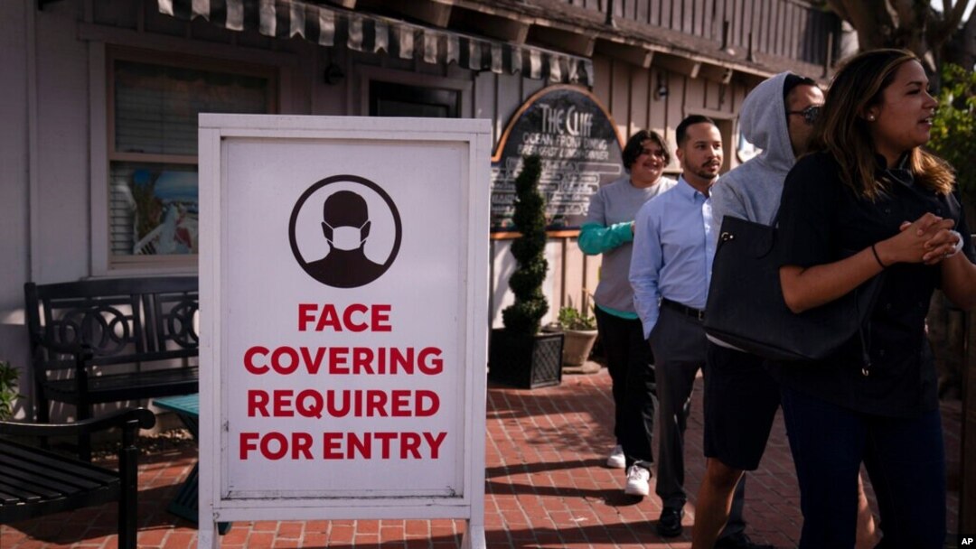 People without masks walk past a sign requesting customers to wear masks in Laguna Beach, Calif., May 17, 2021. 