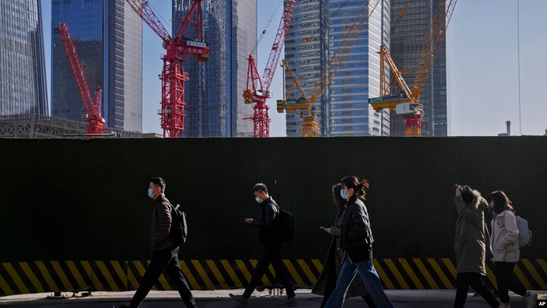 FILE - People wearing face masks walk by construction cranes near office buildings at the central business district in Beijing on March 15, 2023. 